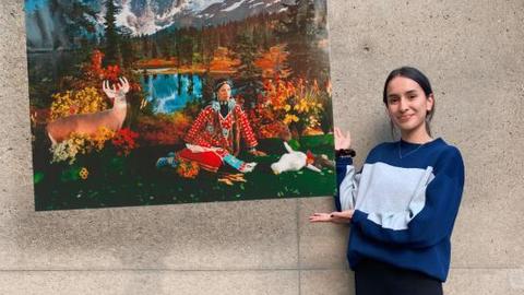 Jacqueline stands against a grey wall. To her left is a large image of the photograph "Indian Summer" by Wendy Red Star. Jacqueline is holding up her hands as if she were holding or presenting the image.