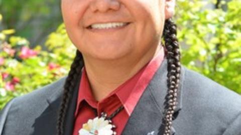 Jason Packineau smiling, hair in two braids, wearing a red button up shirt under a black blazer
