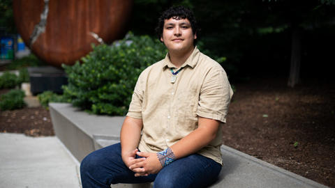 Victor Lopez-Carmen sitting on a concrete bench in a tan shirt with Indigenous jewelry around his neck and on his wrist