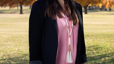 Kelli Mosteller in a pink shirt and black blazer standing in a field in front of trees during the fall