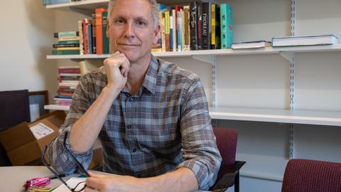 Phil Deloria sitting at a desk in front of bookshelves in a light colored plaid flannel